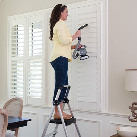 Woman cleaning window blinds while standing on a strong, sturdy step stool